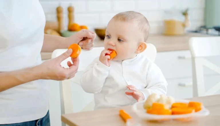 Baby sitting upright in a highchair and eating soft finger foods with an adult nearby