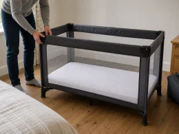 Parent checking the locked frame of an empty travel cot with a firm flat mattress